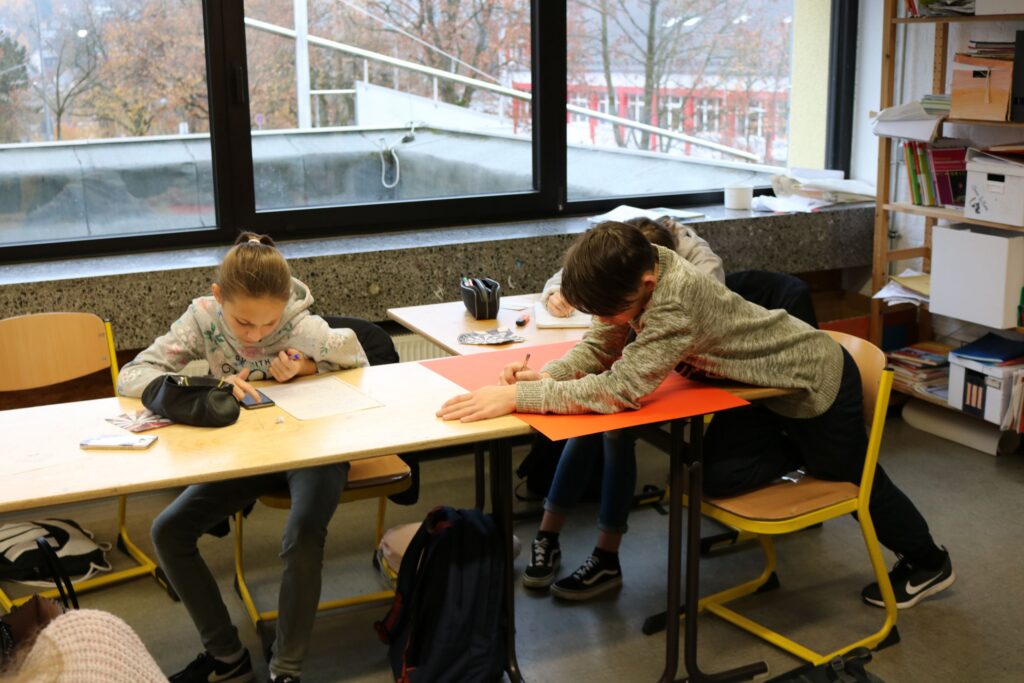 Zwei Schüler arbeiten an Tisch, einer zeichnet, der andere schreibt.