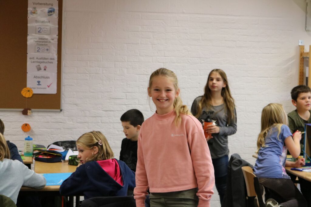 Ein Mädchen lächelt in einem Klassenraum, andere Kinder stehen im Hintergrund.