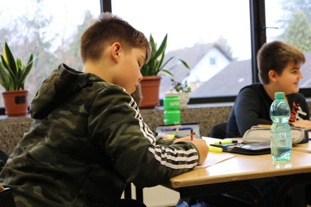 Schüler sitzt am Tisch und schreibt. Im Hintergrund Pflanzen und andere Schüler.