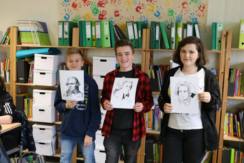 Drei Schüler halten Zeichnungen von berühmten Persönlichkeiten in der Hand.