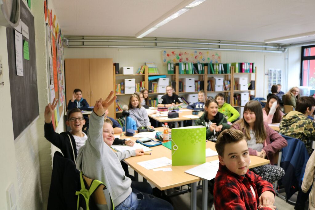 Schüler*innen sitzen in einem Klassenzimmer und schauen freundlich in die Kamera.