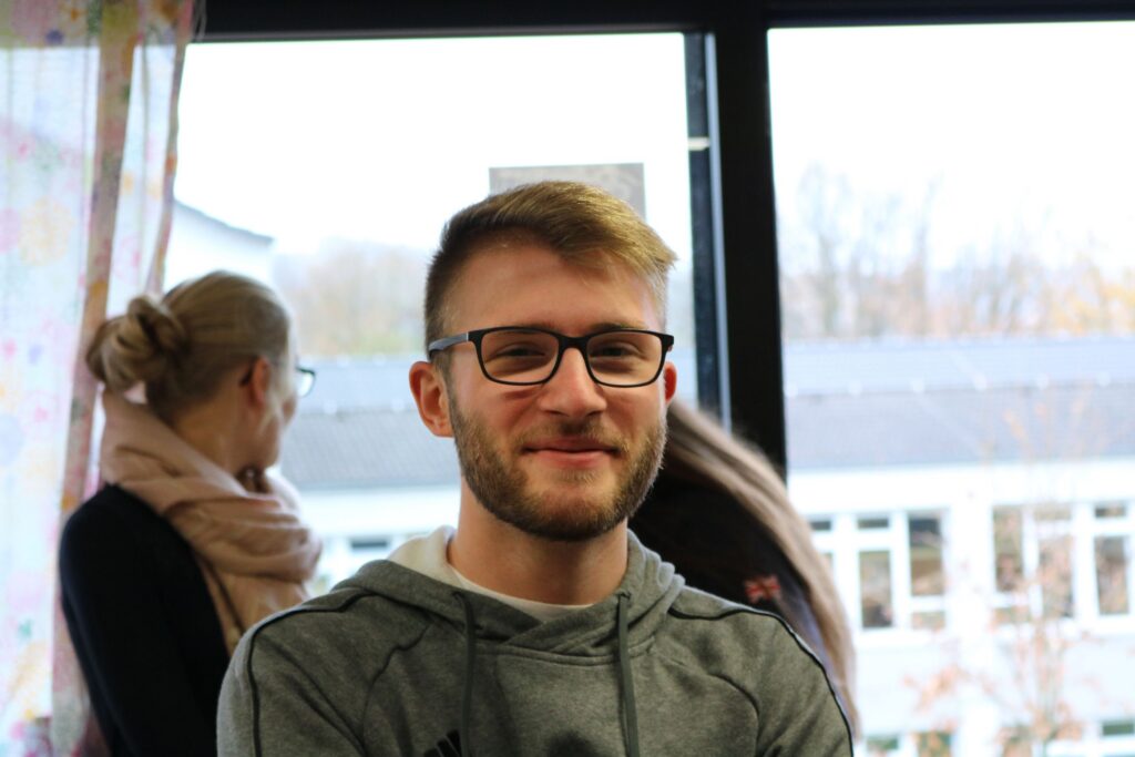 Junger Mann mit Brille lächelt vor einem Fenster, im Hintergrund eine weitere Person.