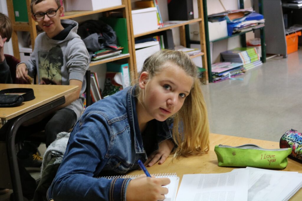 Schülerin schreibt an einem Tisch, im Hintergrund weitere Schüler sichtbar.