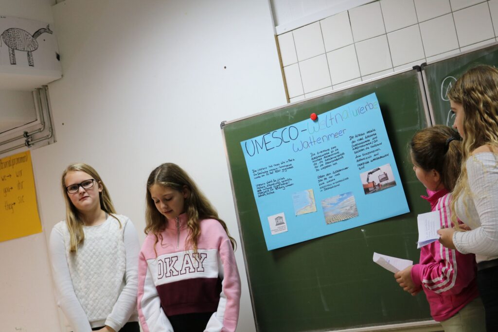 Schülerinnen präsentieren ein Plakat zu UNESCO im Klassenzimmer.