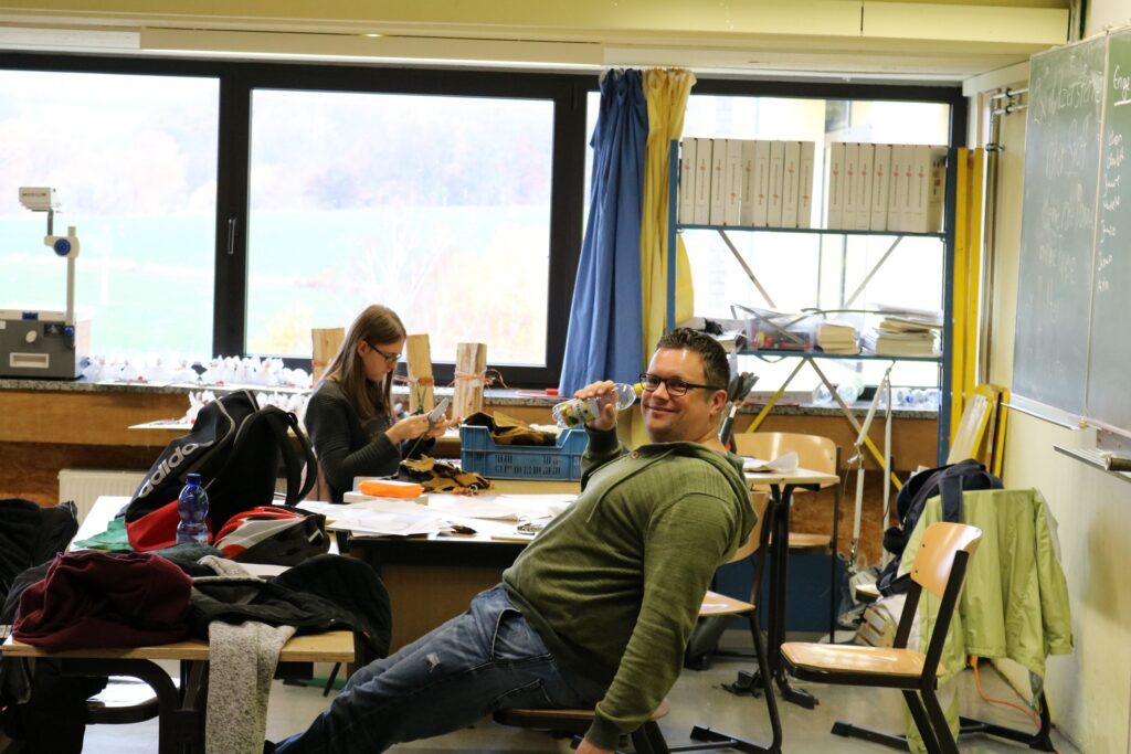 Ein Schüler sitzt lässig auf einem Tisch in einem Klassenzimmer. Im Hintergrund arbeitet eine Mitschülerin.