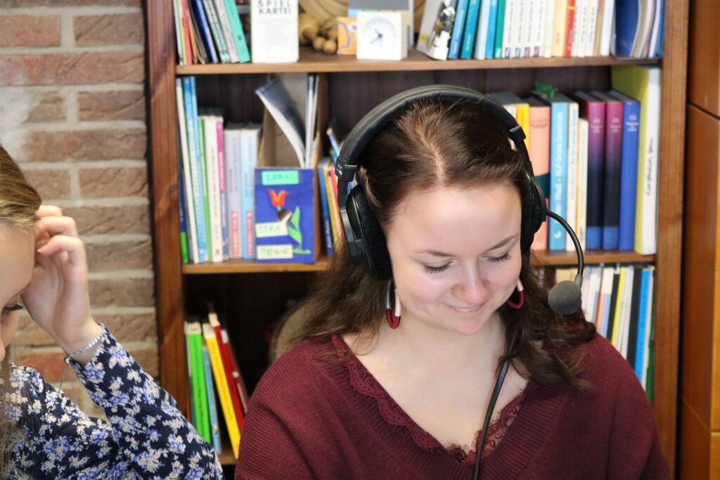 Frau mit Headset sitzt vor einem Bücherregal und lächelt.