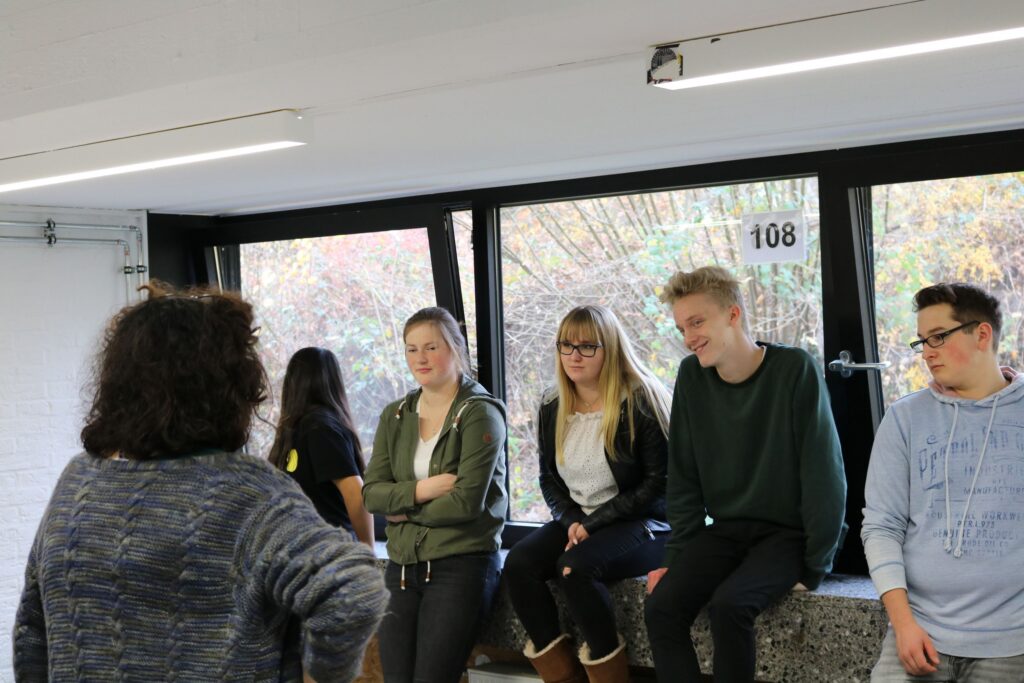 Gruppe von Schülern sitzt am Fenster, eine Lehrkraft steht davor und spricht.