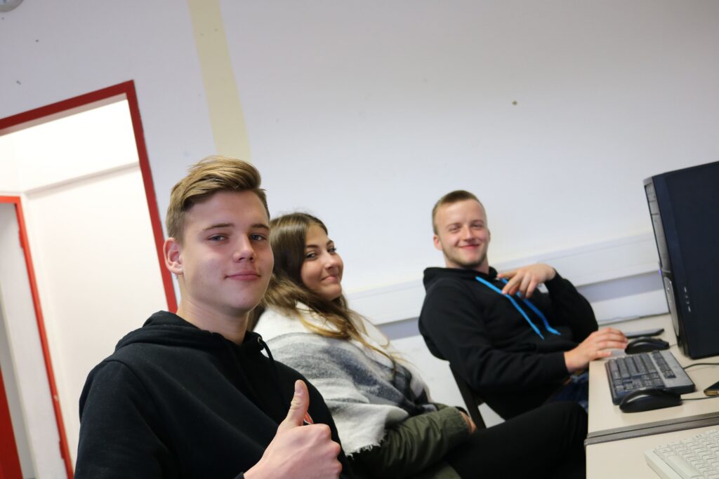 Drei Schüler sitzen an Computern und lächeln in die Kamera.
