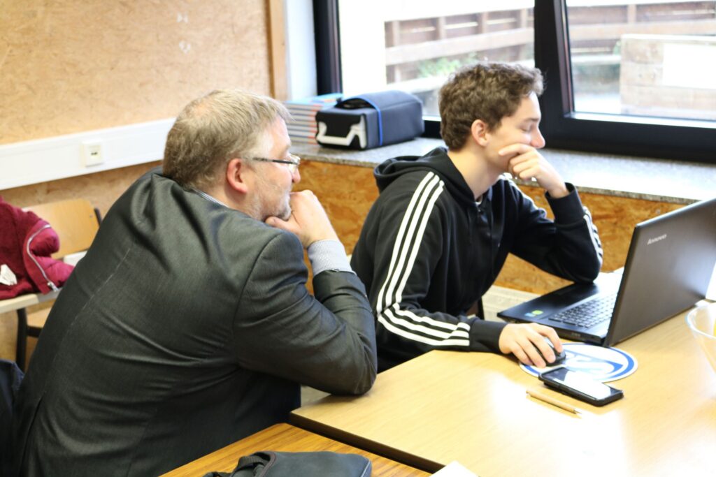 Zwei Personen sitzen an einem Tisch und schauen auf einen Laptop.