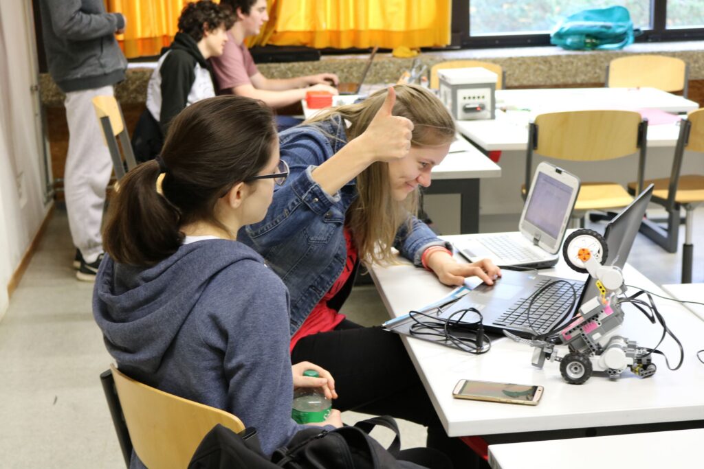 Schülerinnen arbeiten am Computer und einem Roboter-Projekt in einem Klassenzimmer.