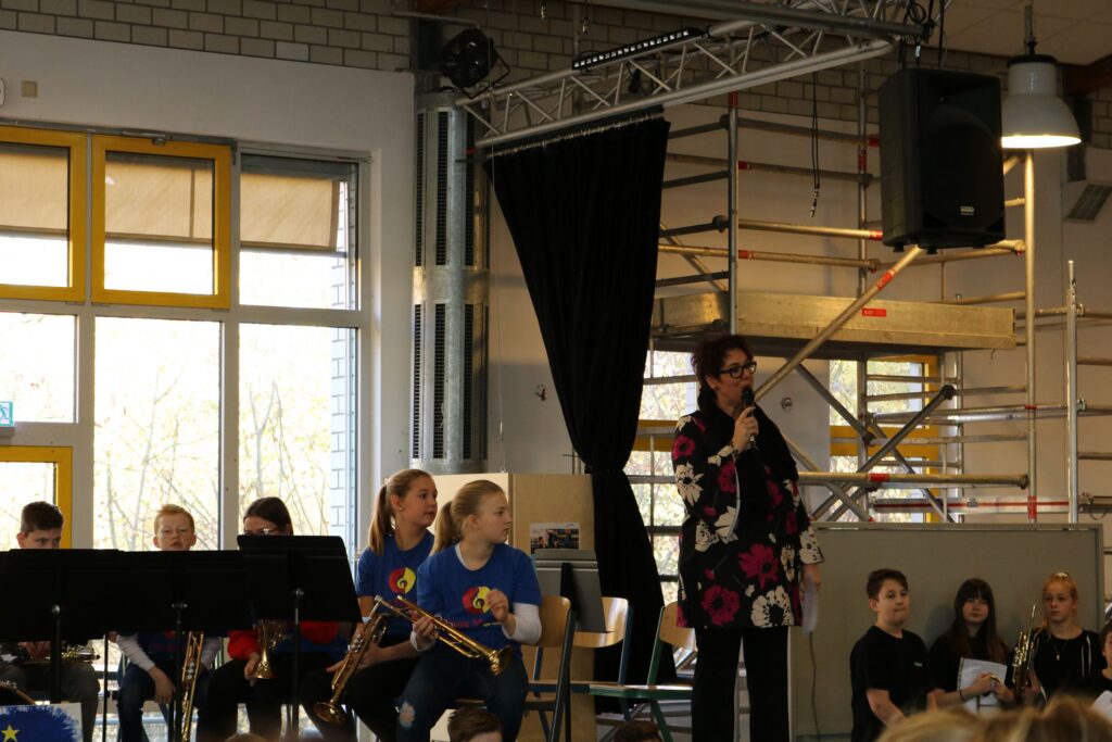 Schüler spielen im Hintergrund Musik, eine Frau spricht im Vordergrund.