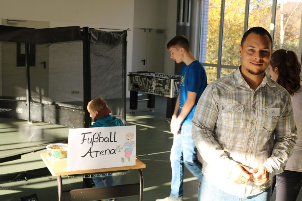 Junge mit einem Schild "Fußball Arena" stehen an einem Tisch, im Hintergrund sind weitere Personen.