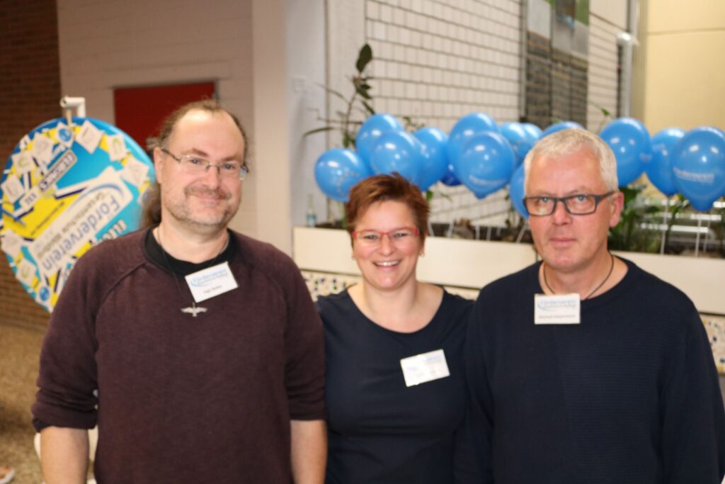 Drei Personen stehen vor einem Hintergrund mit blauen Luftballons.