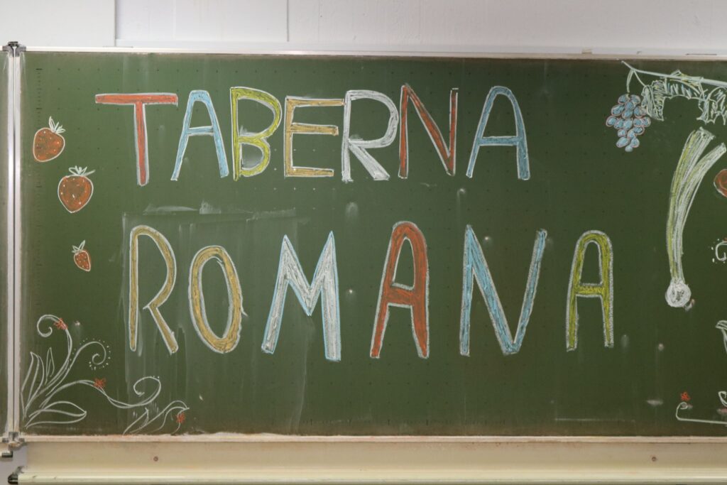 Bunte Schriftzüge "TABERNA ROMANA" auf einer grünen Schultafel.