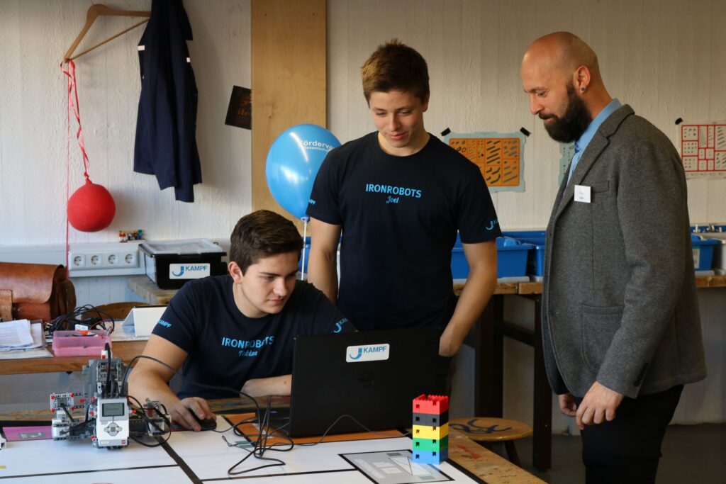 Drei Personen arbeiten an einem Computer mit Robotertechnik in einem Klassenraum.
