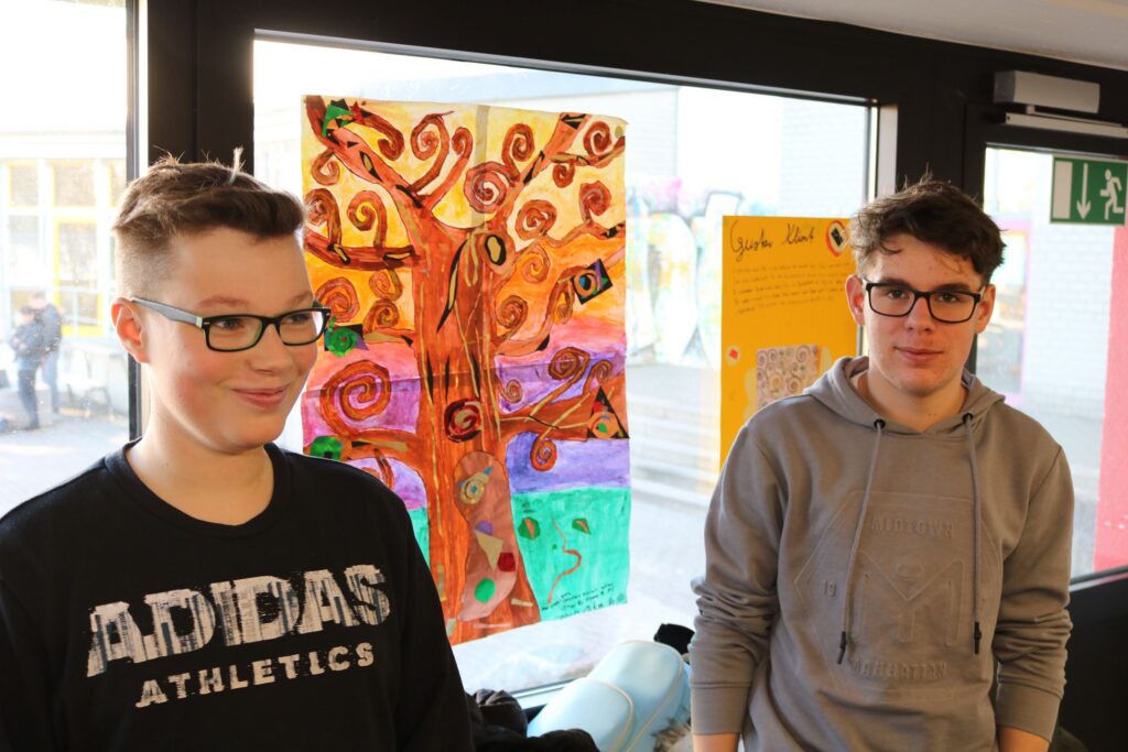 Zwei Schüler stehen vor einem bemalten Fenster mit einem Baum-Motiv.