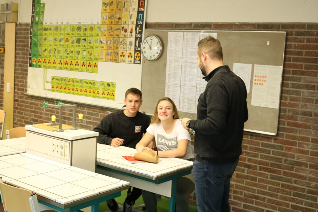 Zwei Schüler sitzen an einem Tisch, ein Lehrer spricht mit ihnen.
