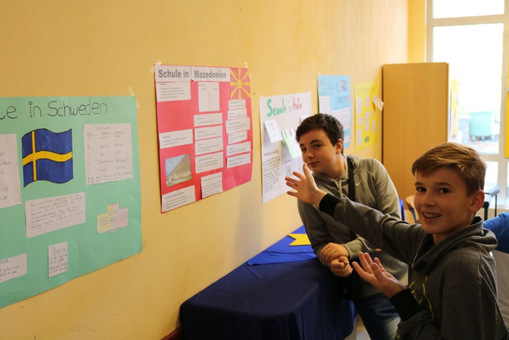 Zwei Schüler präsentieren an einer Wand Plakate zu Schweden und ihrer Schule.