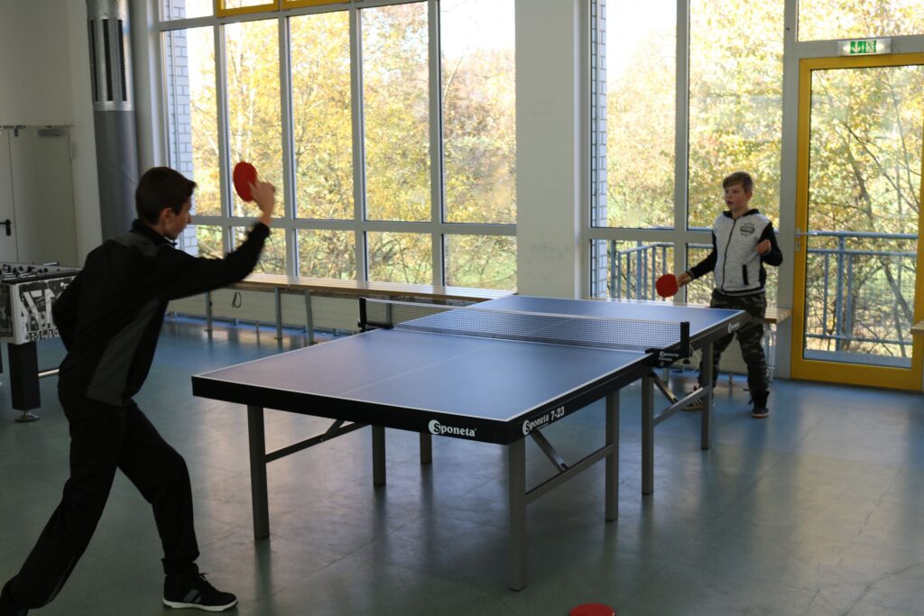 Zwei Schüler spielen Tischtennis in einem hellen Raum mit großen Fenstern.