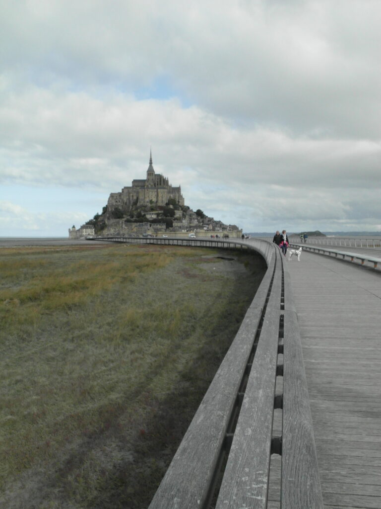 Blick auf Mont Saint-Michel mit Holzsteg und Landschaft bei bewölktem Himmel.
