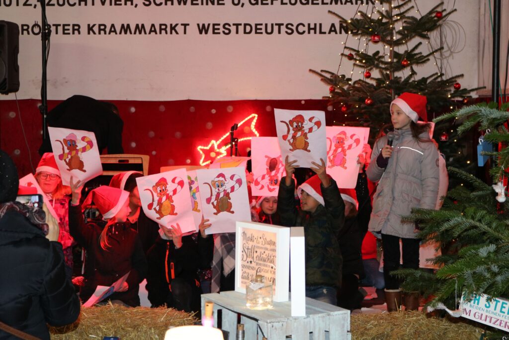 Kinder in Weihnachtsmützen präsentieren Zeichnungen auf einer Bühne. Weihnachtsbaum im Hintergrund.