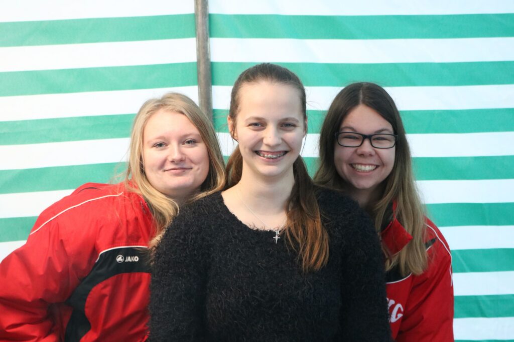 Drei Frauen lächeln vor einem gestreiften Hintergrund.