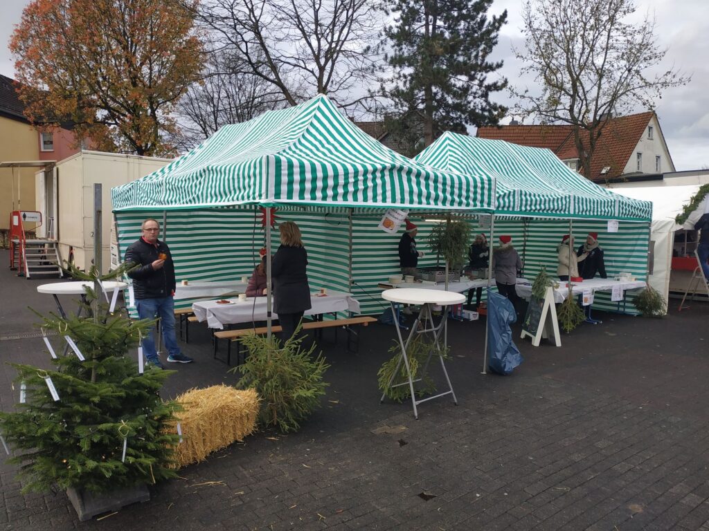 Marktstände mit grün-weißen Streifen, wenige Personen stehen dabei. Herbstliche Umgebung.