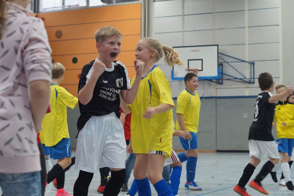 Kinder in Sportkleidung jubeln und feiern in einer Sporthalle.