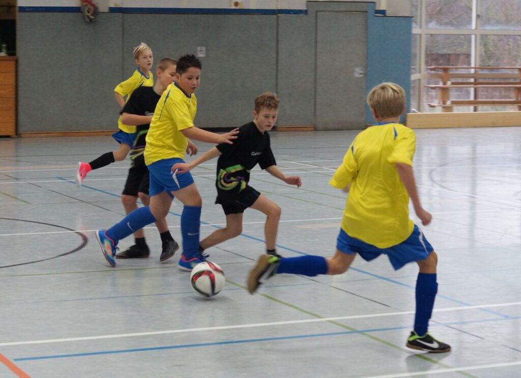 Kinder spielen in einer Halle Fußball, zwei tragen gelbe Trikots.