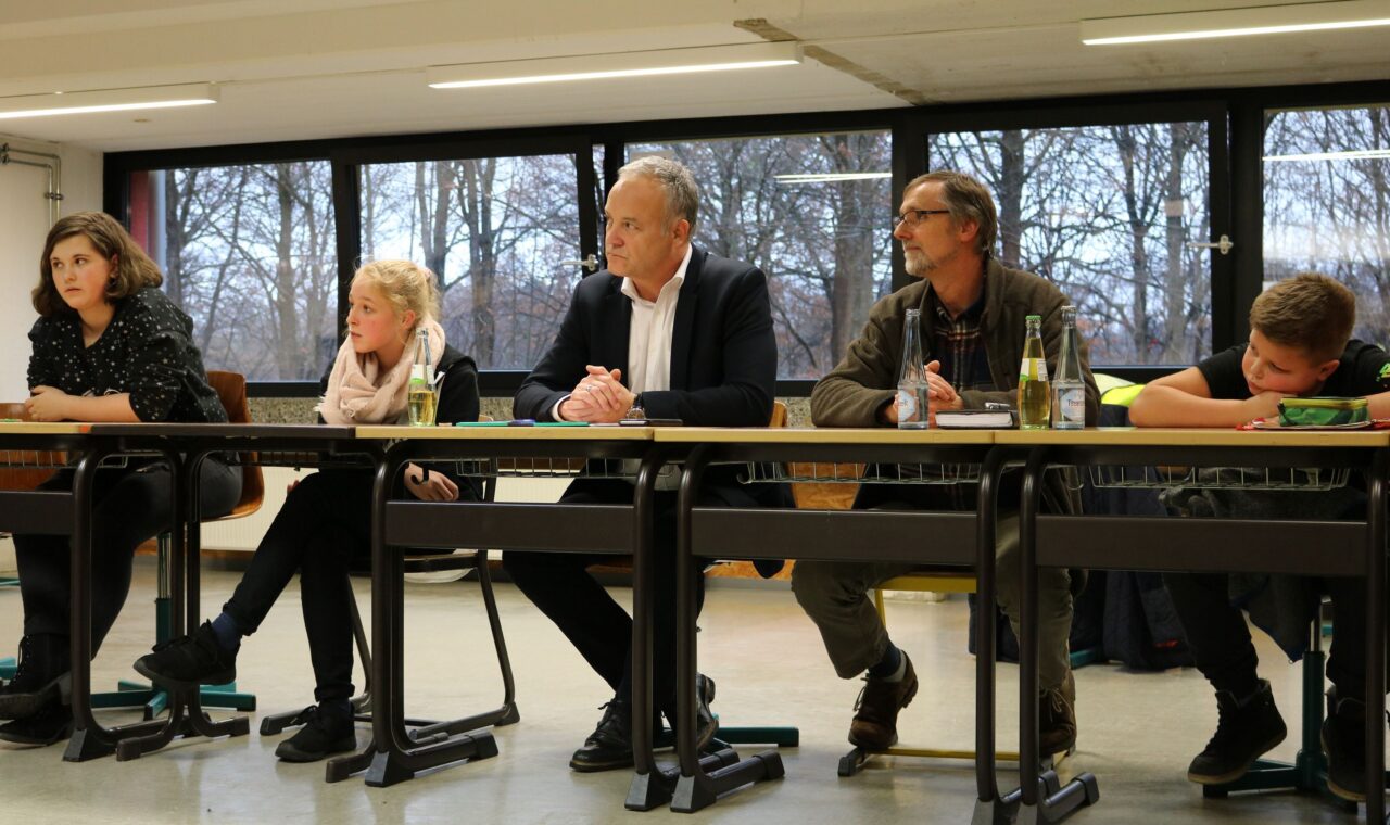 Fünf Personen sitzen an einem Tisch in einem Klassenraum.