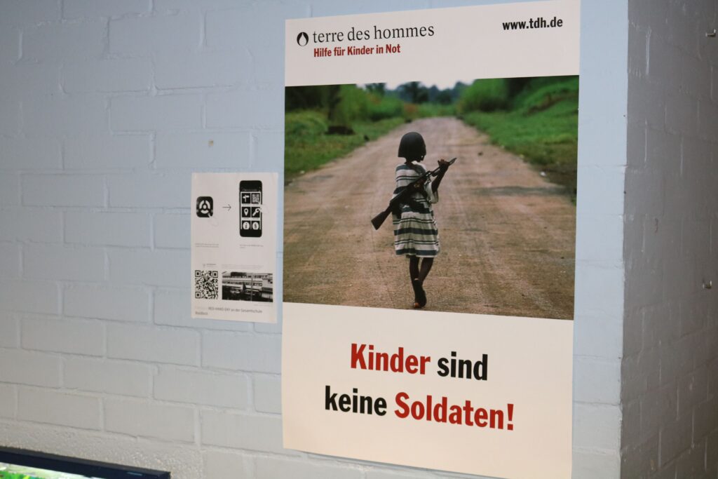 Ein Plakat mit dem Text "Kinder sind keine Soldaten!" und einem Kind auf einem Weg.