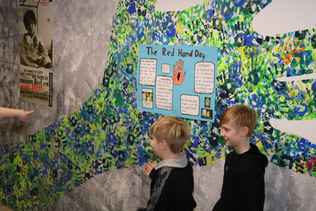Zwei Kinder stehen vor einer bunten Wand mit Plakaten und Infos zum Red Hand Day.