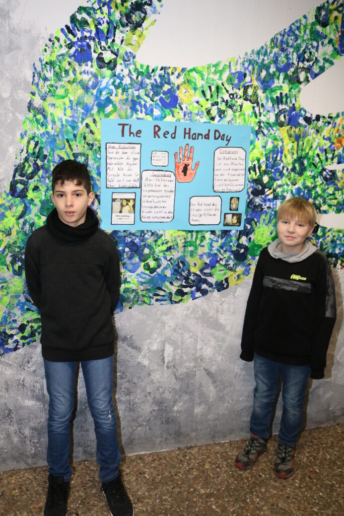 Zwei Kinder stehen vor einer Wand mit einem Plakat zum "Red Hand Day".