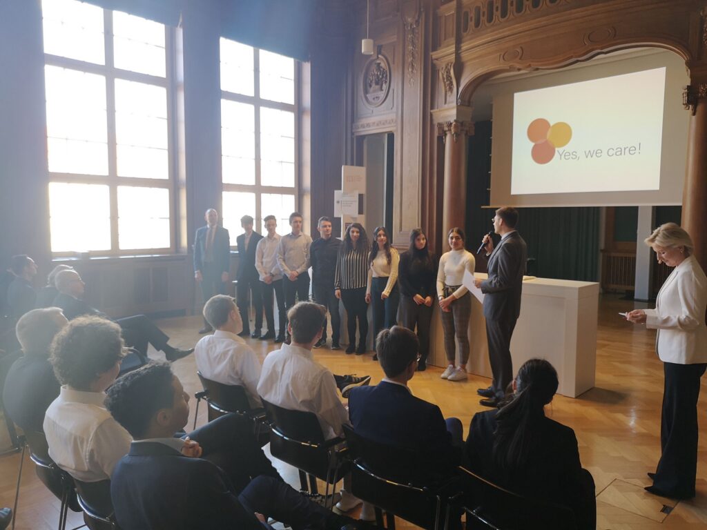 Schüler stehen auf der Bühne, Zuschauer sitzen davor in einem Saal.