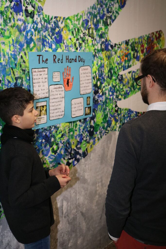Schüler präsentiert ein Plakat zum Red Hand Day vor einer bunten Wand.
