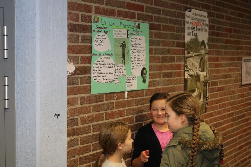 Drei Kinder stehen vor einem Plakat an einer Ziegelwand in einem Flur.