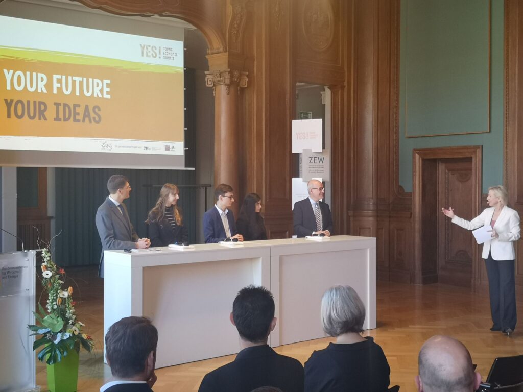 Diskussionsrunde mit vier Personen am Tisch und einer Rednerin im Vordergrund.