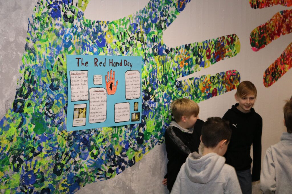 Bunte Hände an der Wand mit einem Plakat zum "Red Hand Day" und Kindern im Gespräch.