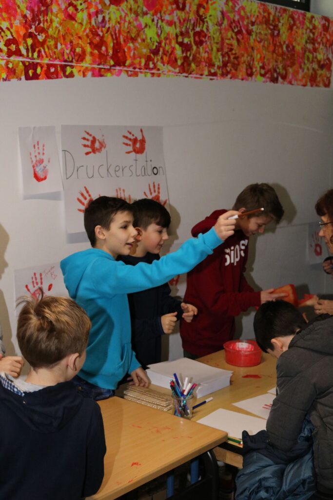 Kinder arbeiten an einem Tisch mit Bastelmaterialien und Handabdrücken an der Wand.