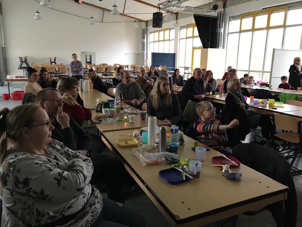 Eltern und Schüler sitzen in einem Klassenraum und verfolgen eine Präsentation.