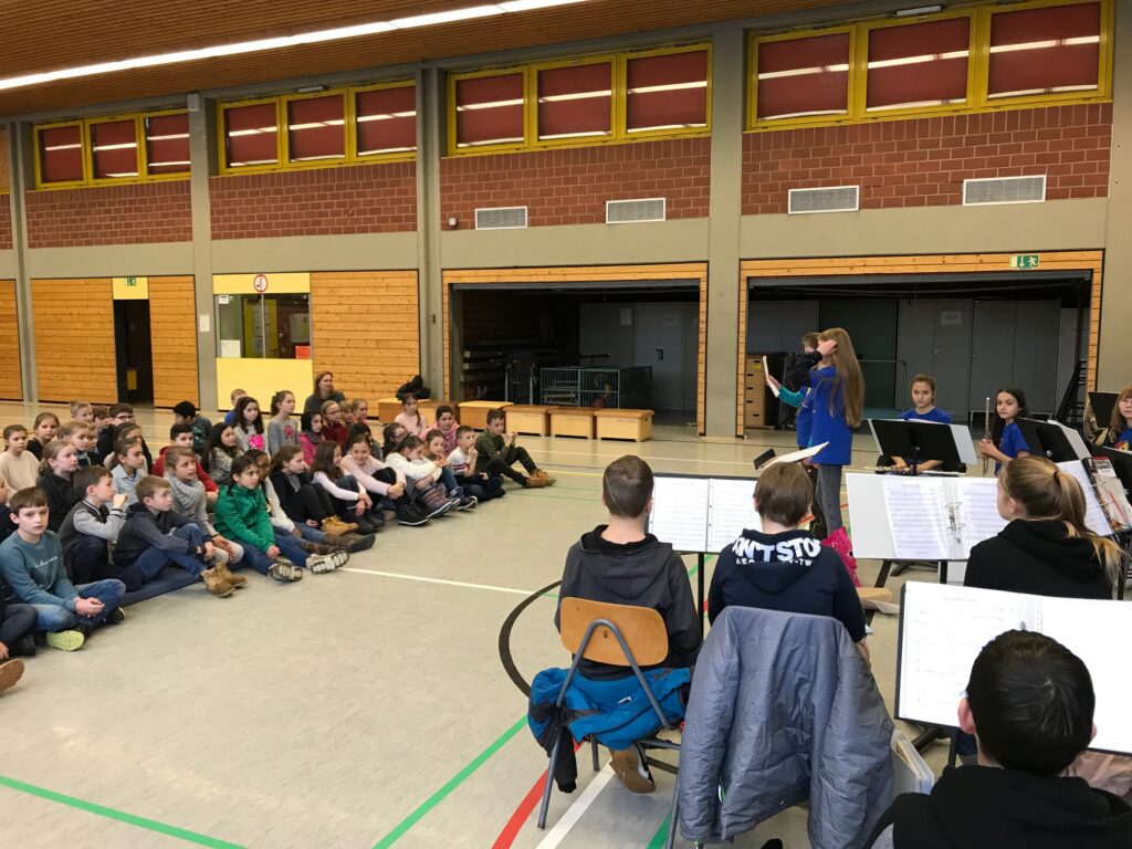 Schüler sitzen auf dem Boden und schauen auf eine Aufführung in einer Sporthalle.