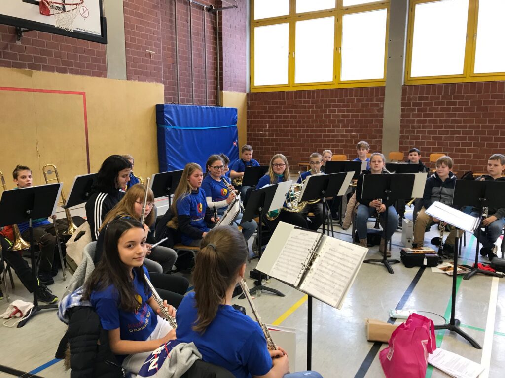 Schülerinnen und Schüler einer Musikgruppe sitzen mit Instrumenten in einer Turnhalle.