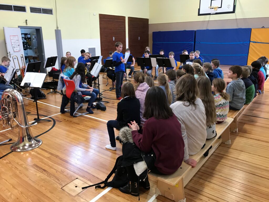 Schüler sitzen auf Bänken und beobachten eine Musikprobe in der Turnhalle.