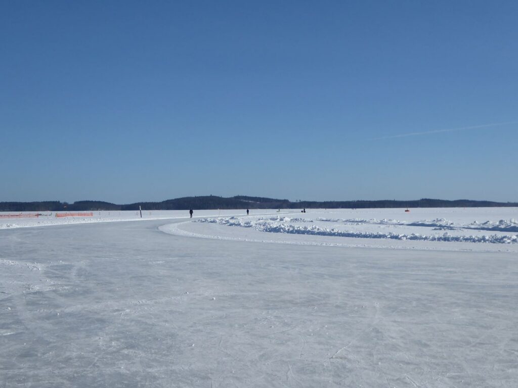 Eisfläche mit Schlittschuhfahrern und Berglandschaft im Hintergrund.