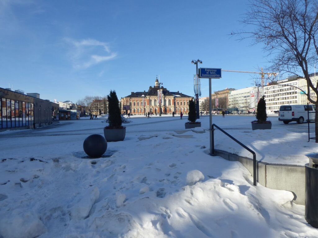 Schneebedeckter Platz mit historischem Gebäude im Hintergrund und klar blauem Himmel.