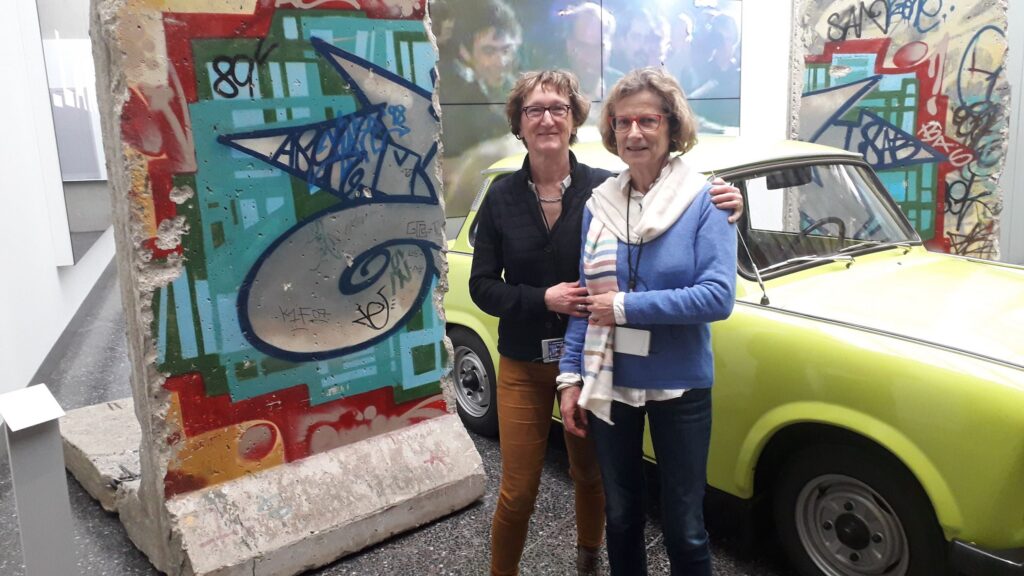 Zwei Frauen stehen vor einem graffitibemalten Mauerstück und einem oldtimer Auto.