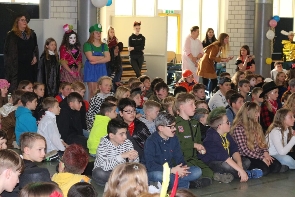 Schüler in Kostümen sitzen und stehen in einer Aula während einer Veranstaltung.