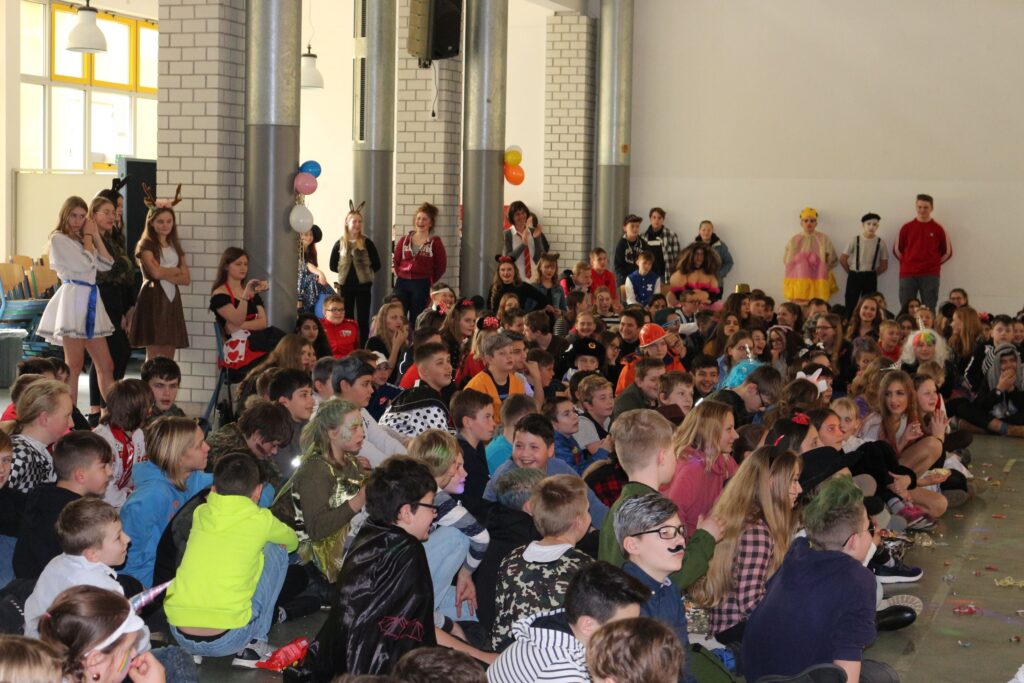 Vielzahl von Kindern und Jugendlichen sitzt in einer Halle und schaut auf die Bühne.