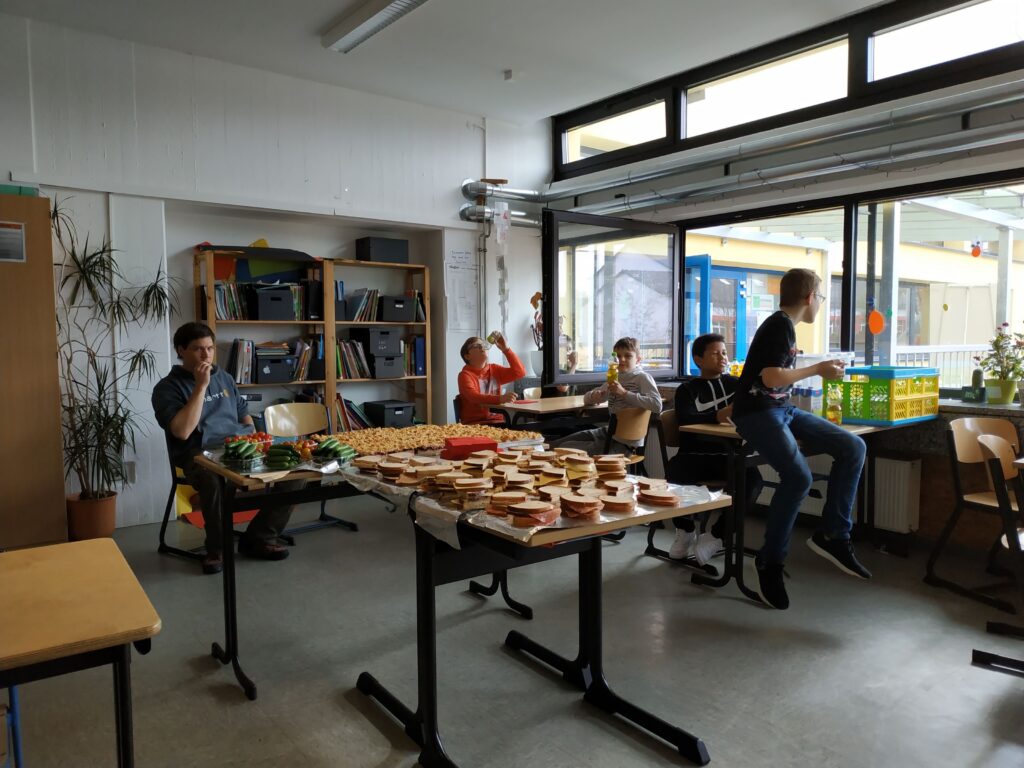 Schüler sitzen in einem Klassenraum, essen Snacks und arbeiten an Tischen.