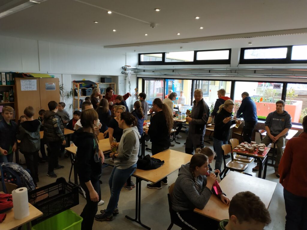 Viele Schüler und Erwachsene in einem Klassenraum bei Aktivitäten. Tische sind voll.